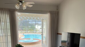 A living room with a TV, a coffee table with a plant, a sofa, a ceiling fan, and a view of a swimming pool through a glass door. - Quiet Private Guest House (North Fort Myers)