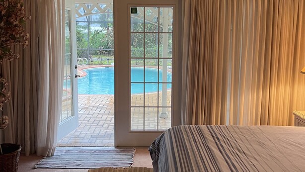 A bedroom with a bed, a ceiling fan, a view of a swimming pool, and a wicker chair. - Quiet Private Guest House (North Fort Myers)