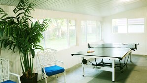 Mesa de pingue-pongue em um quarto com paredes brancas e janelas grandes.