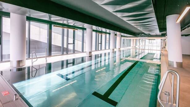 En modern inomhuspool med väggar av genomskinligt glas, metallräcken och ett grönt golv.