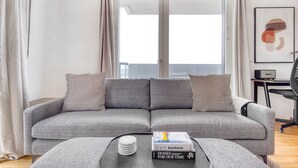 Ett modernt vardagsrum med en grå soffa, mönstrade kuddar, ett soffbord med böcker och ett stort fönster med gardiner.