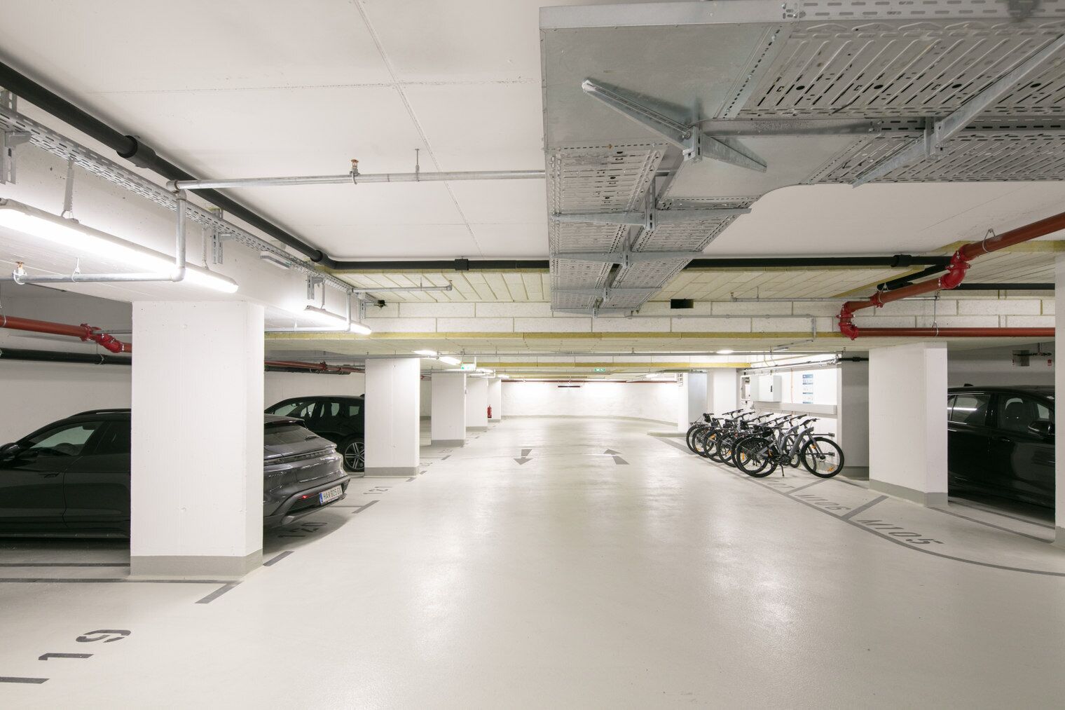 Un parking à plusieurs niveaux aux murs et au sol blancs, où l’on peut voir plusieurs voitures et vélos garés.