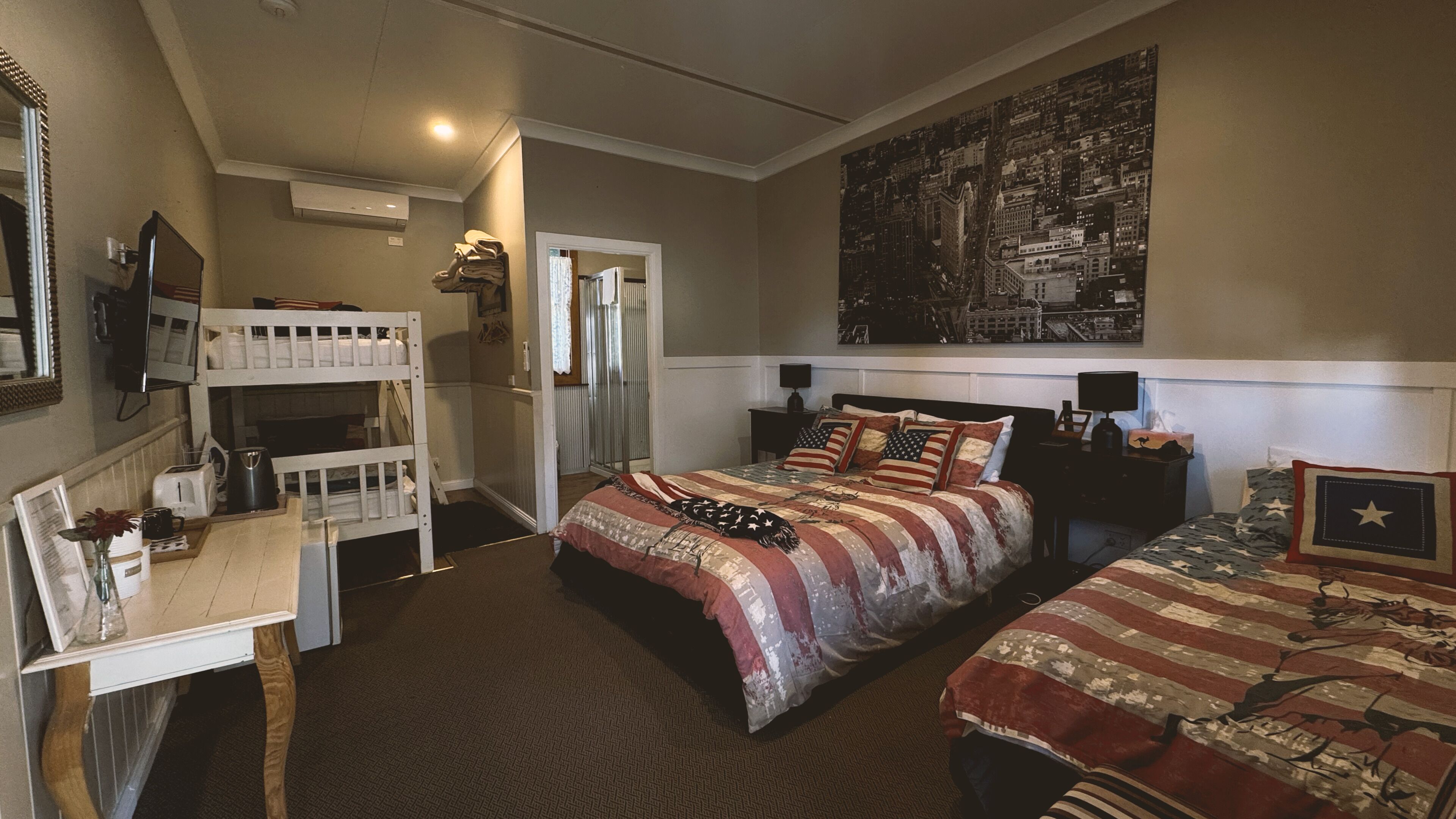 Um quarto com beliches, uma escrivaninha, uma cadeira, uma cama com colcha temática da bandeira americana e uma mesa de cabeceira com abajur, além de uma pintura da paisagem urbana na parede.