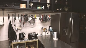 Uma cozinha com geladeira de aço inoxidável, um suporte para utensílios pendurado, cafeteira e chaleira sobre a bancada.