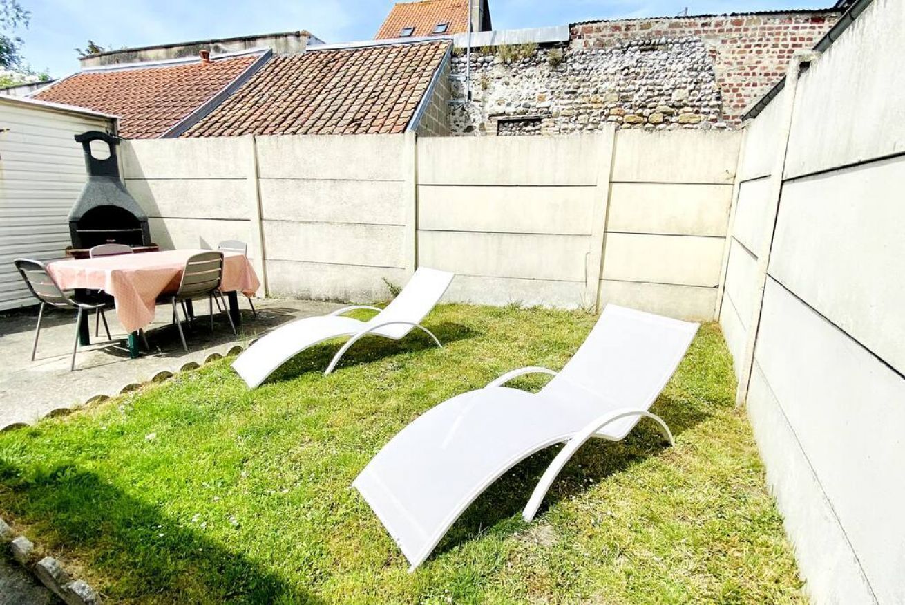 Un patio trasero con una parrilla, una mesa con sillas y dos sillones blancos.