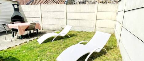 Un patio trasero con una parrilla, una mesa con sillas y dos sillones blancos.