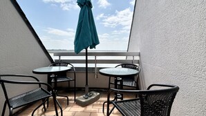 Un balcon avec deux tables rondes et quatre chaises, un parasol de couleur turquoise et une vue sur le ciel et l’eau.