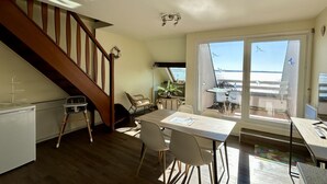 Une pièce avec un escalier en bois, un coin repas avec une table et des chaises, un petit bureau blanc et une grande porte-fenêtre coulissante donnant sur un balcon extérieur avec vue.