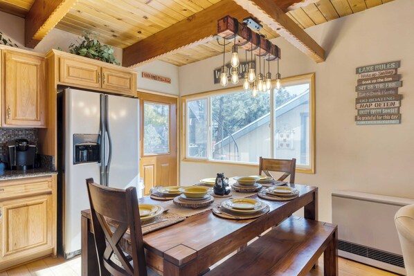 A kitchen with wooden cabinets, a stainless steel refrigerator, and a dining table set with plates and a coffee maker. - 50 Yards to Lake, Boulder Bay Pool Table Lakeside in Bearadise by Avantstay (Big Bear Lake)