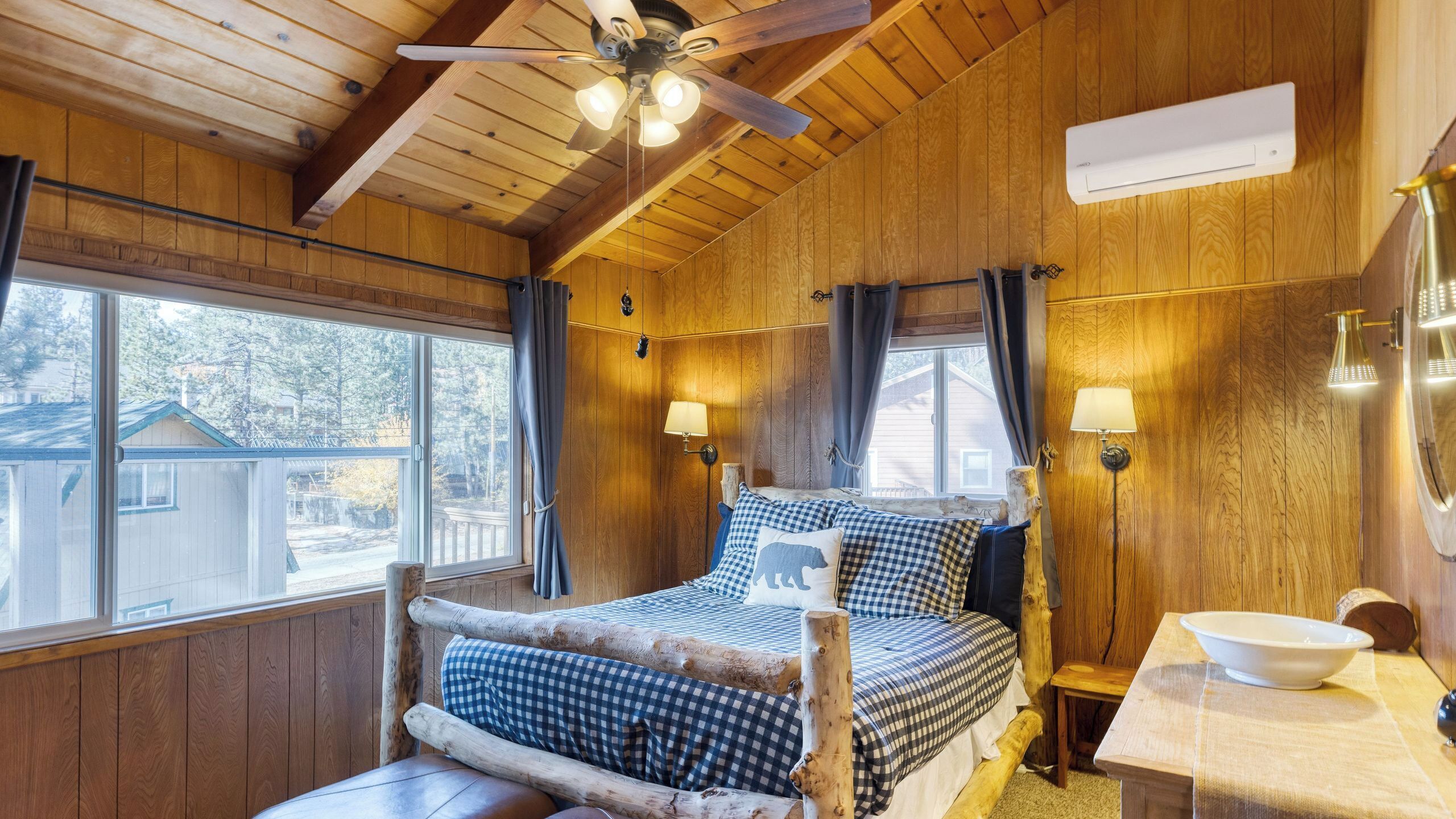 A wooden cabin room with a bed, two wall-mounted lamps, a ceiling fan, and a window with curtains.