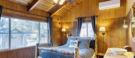 A wooden cabin room with a bed, two wall-mounted lamps, a ceiling fan, and a window with curtains.