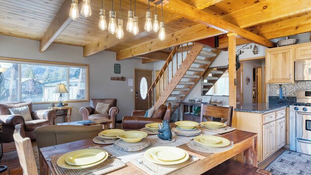 Ein gemütliches Wohnzimmer mit einem Holzespeisetisch, der für eine Mahlzeit gedeckt ist, einer Treppe, die zu einer anderen Ebene führt, und einer Küchenzeile mit Holzschränken und einem Herd.
