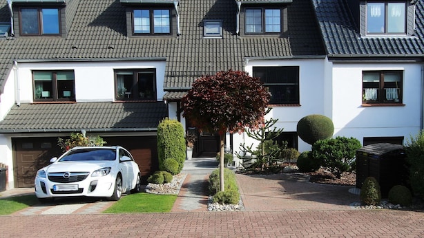 Una casa a due piani con un'auto nel vialetto, un giardino curato e un cielo sereno.