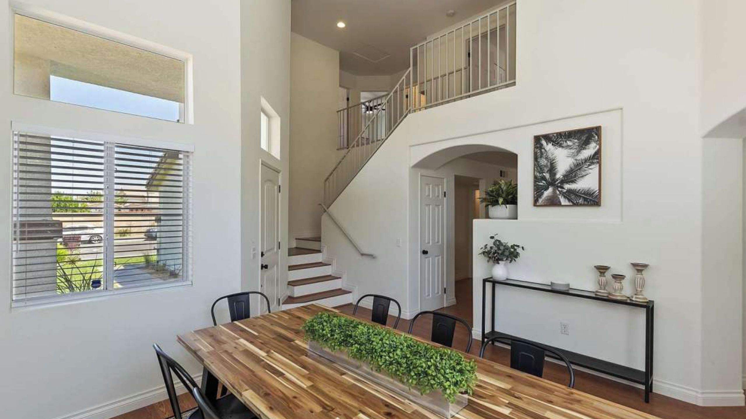 Un comedor amplio con una mesa de madera, sillas y una planta en el centro. Una escalera conduce a un nivel superior con barandilla.