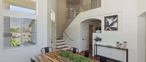 A spacious dining area with a wooden table, chairs, and a central plant. A staircase leads to an upper level with a railing.
