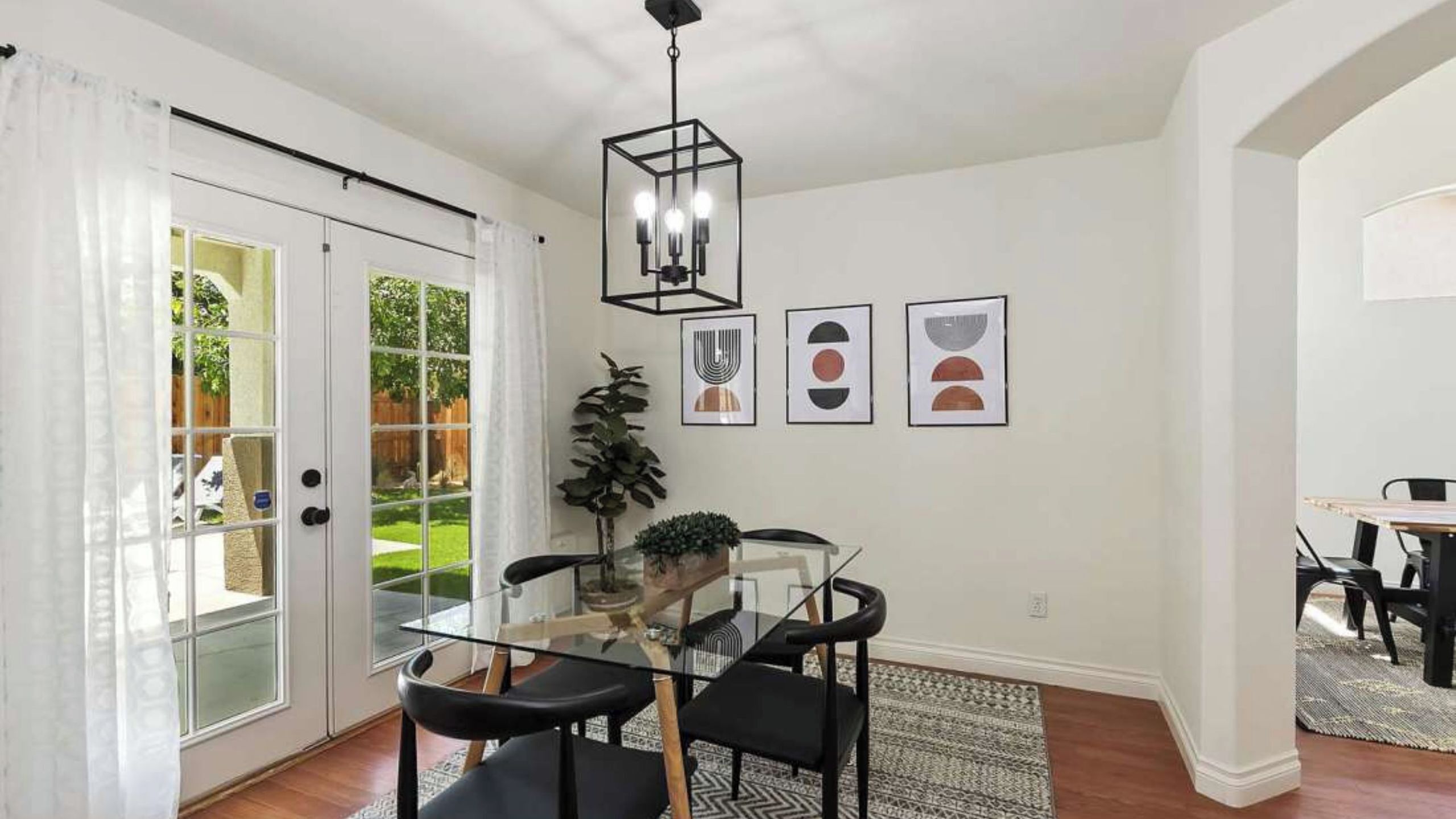 A dining area with a glass table, black chairs, a hanging light fixture, and framed artwork on the wall.