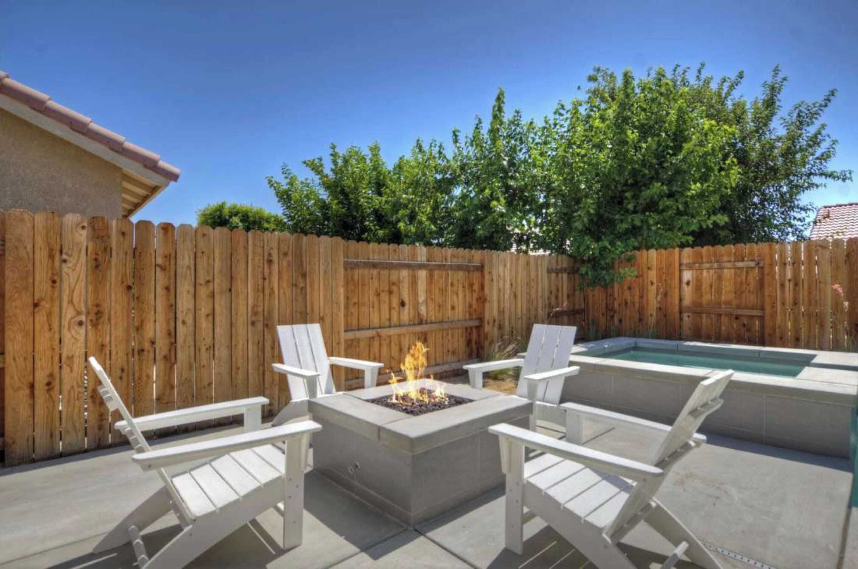 Un patio trasero con cerca de madera, un brasero, sillas blancas para lounge y una piscina.