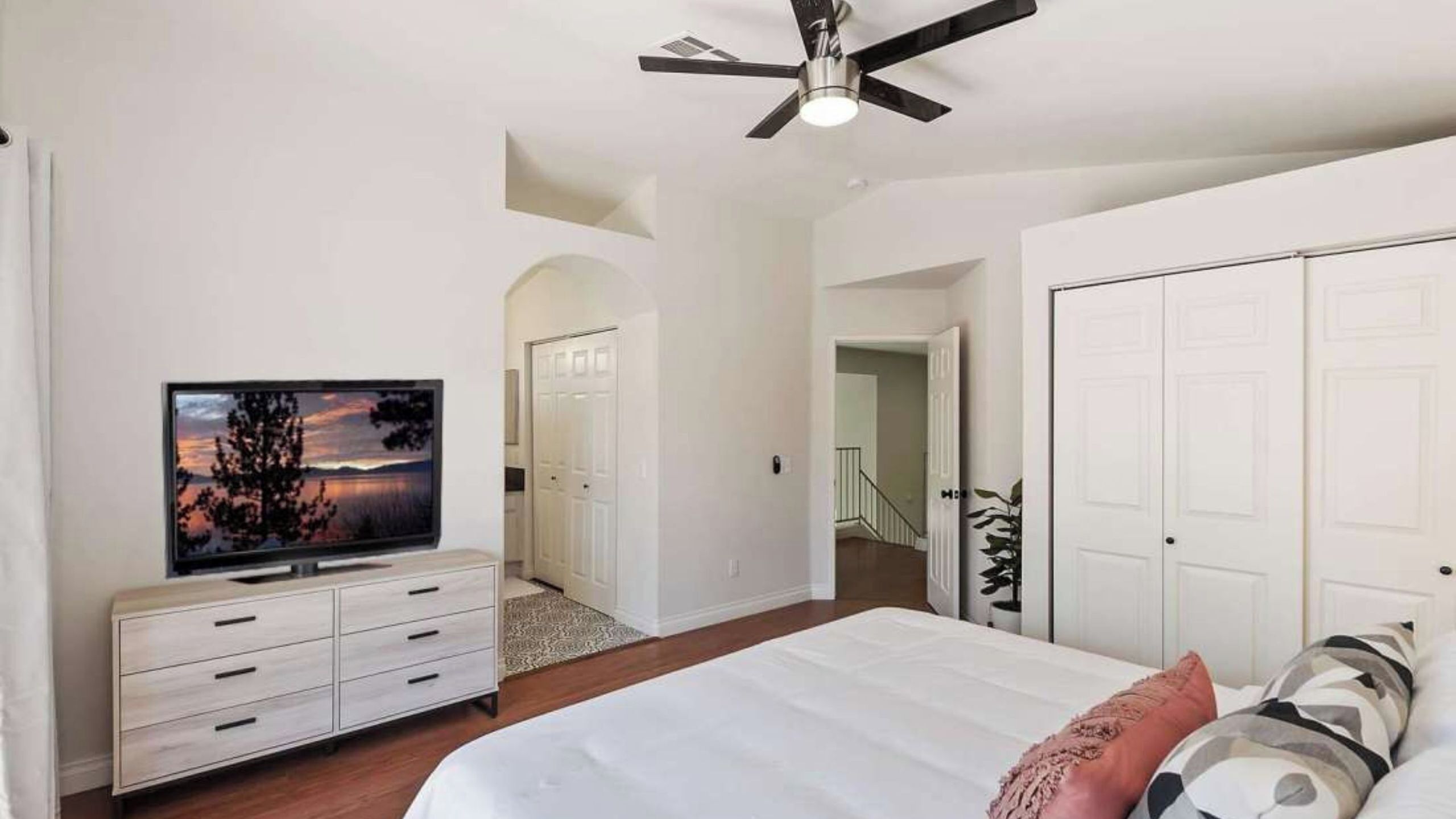 A bedroom with a bed, a TV, a ceiling fan, and a staircase leading to another room.