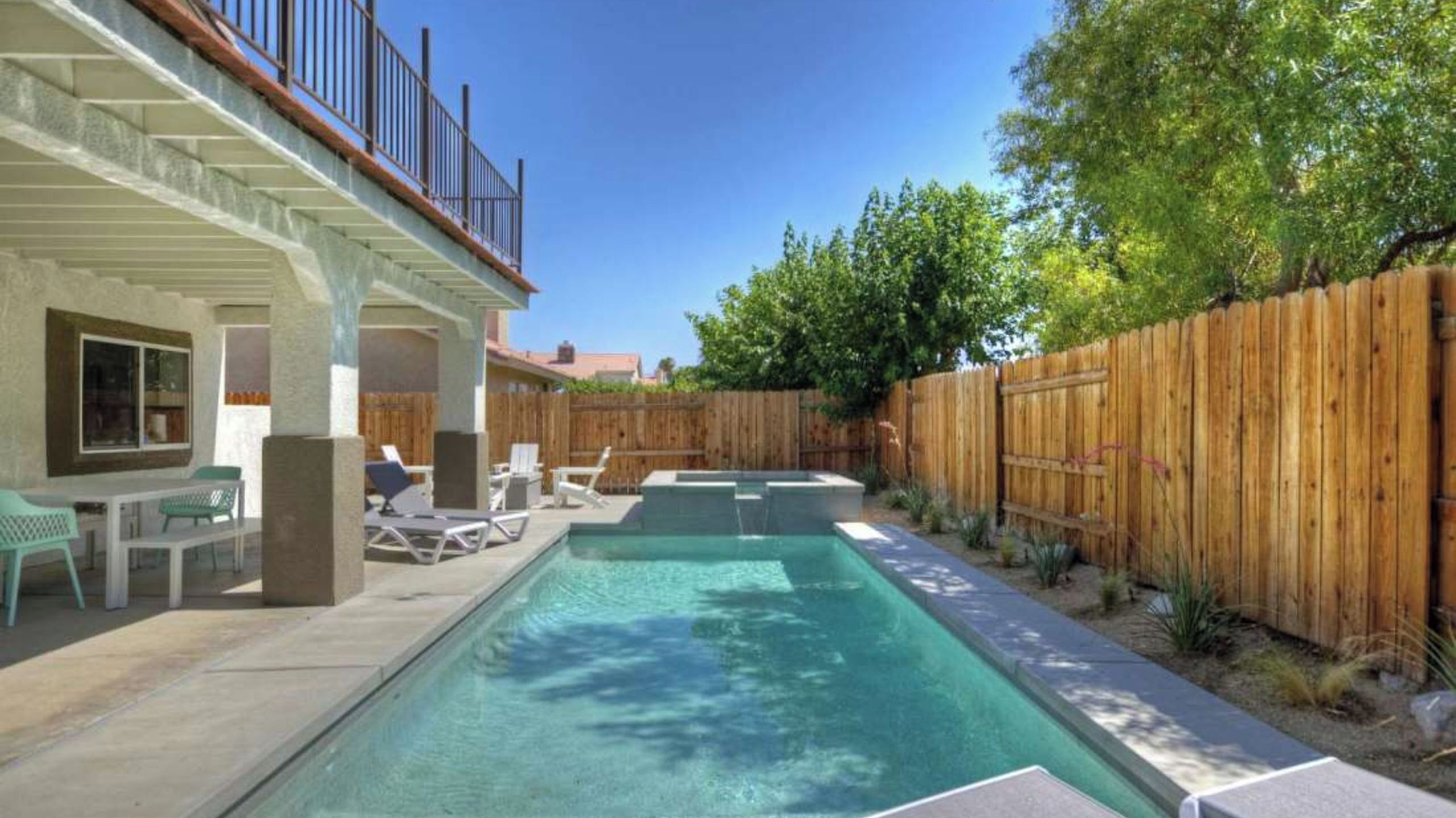 A rectangular swimming pool with a wooden deck, a covered patio area, and a wooden fence.