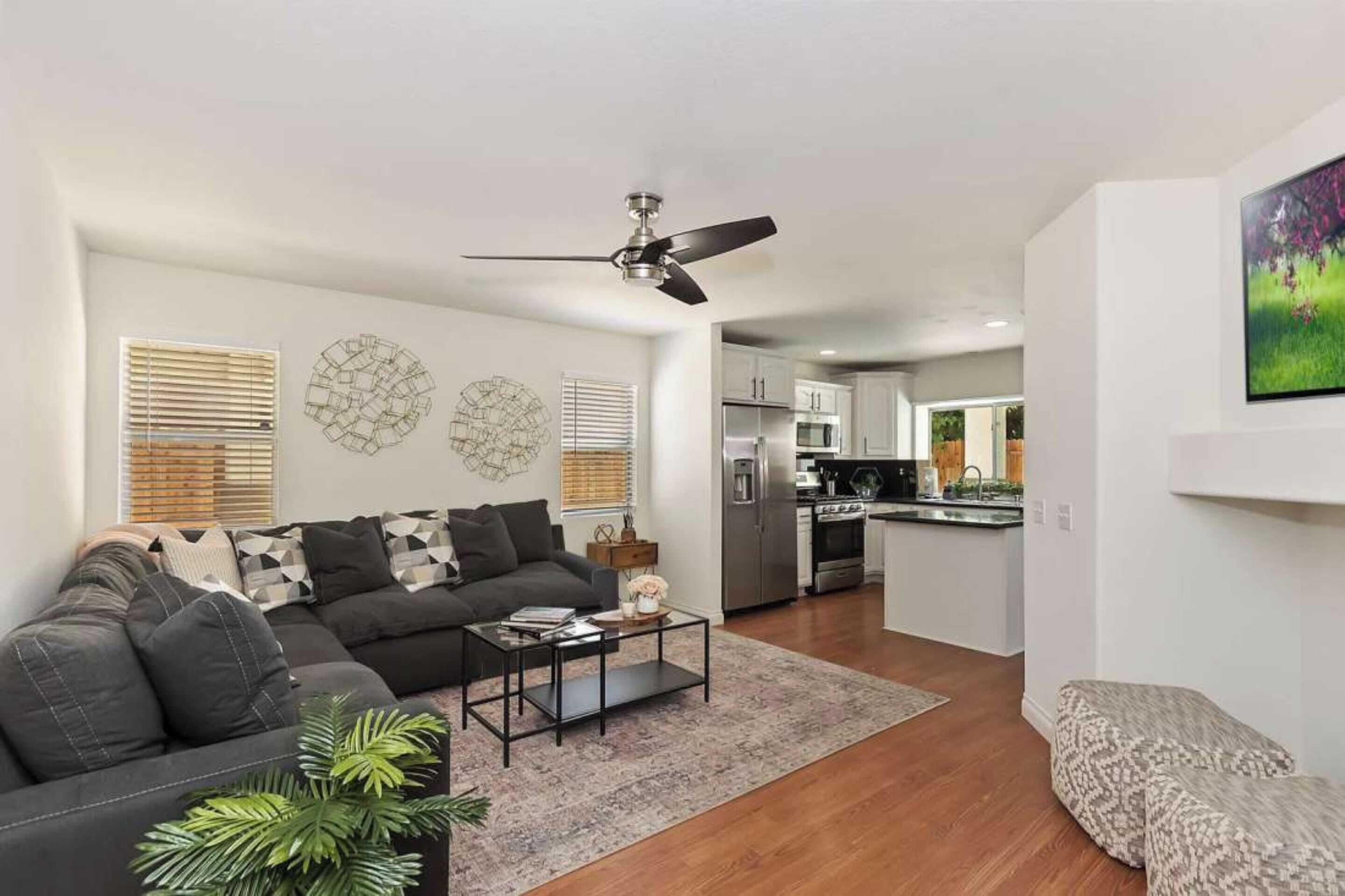 A modern living room with a sectional sofa, a coffee table, and a ceiling fan. There is a kitchen in the background with stainless steel appliances.