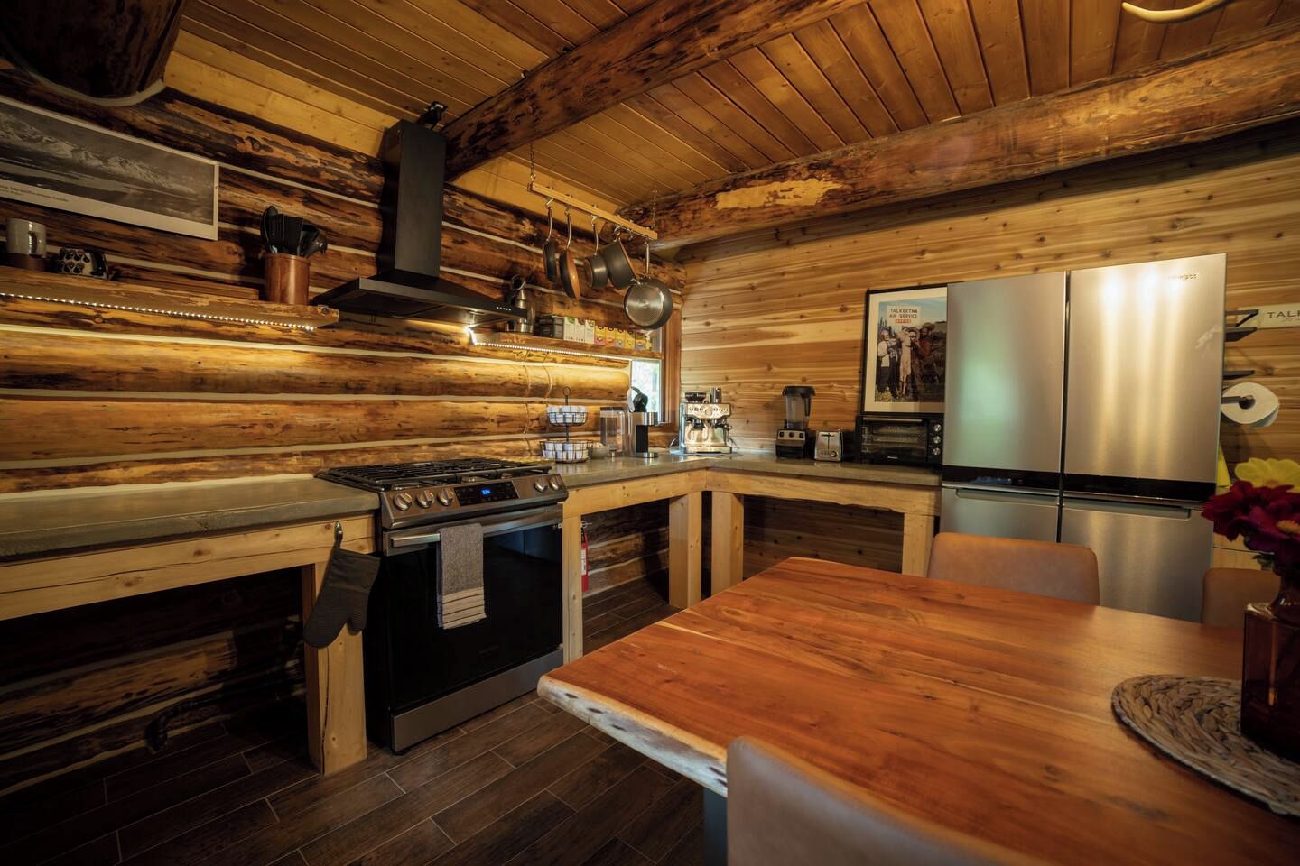 A rustic kitchen with wooden walls, a gas stove, a stainless steel refrigerator, and a wooden dining table.