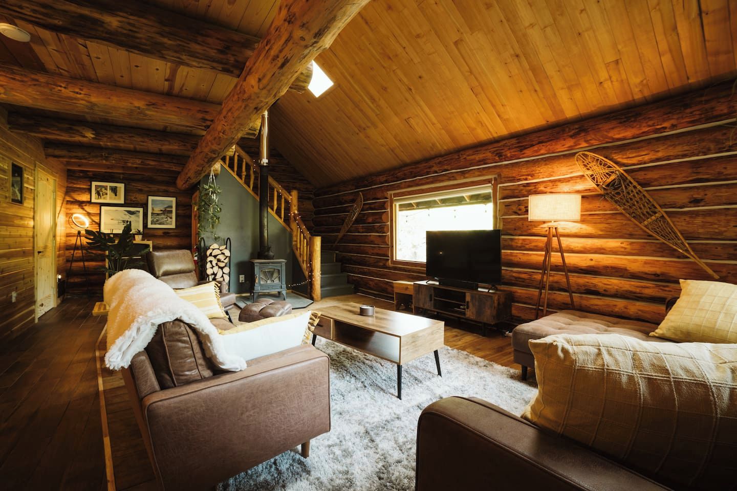 A log cabin interior with a fireplace, wooden walls, a staircase, a television, and a cozy seating area.