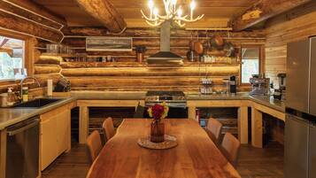 A log cabin-style kitchen with a wooden dining table, chairs, a chandelier, and various kitchen appliances.