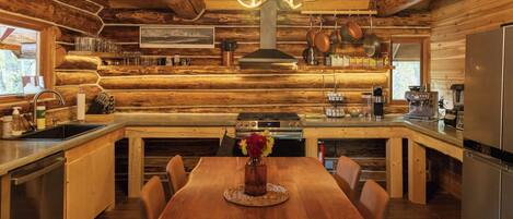 A log cabin-style kitchen with a wooden dining table, chairs, a chandelier, and various kitchen appliances.