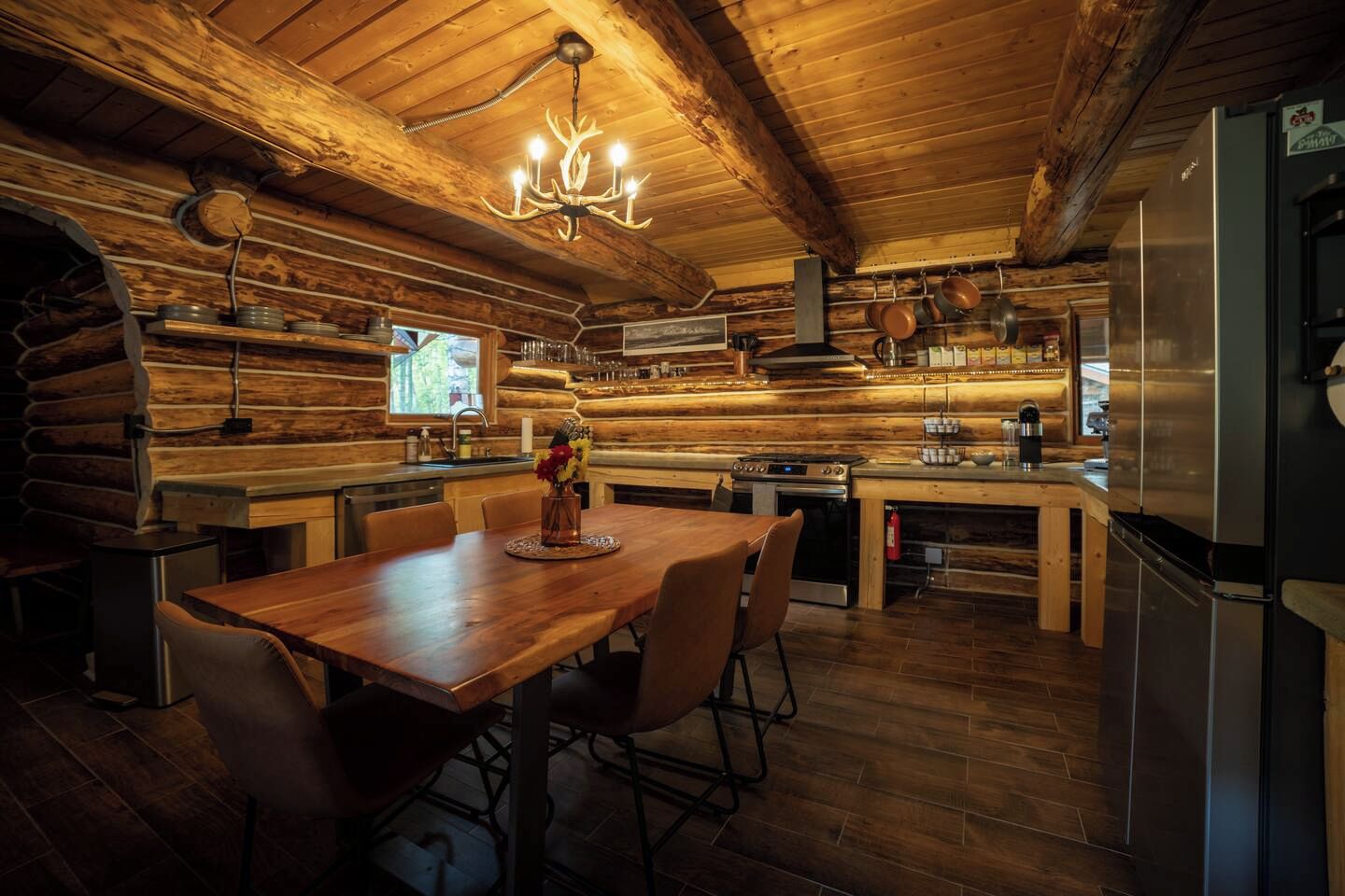Een blokhut interieur met een houten eettafel, stoelen, een kroonluchter, een keuken met moderne apparatuur en een open haard.