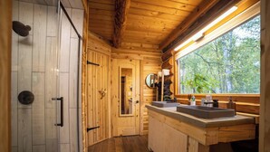 A wooden cabin bathroom with a shower, two sinks, a mirror, and a window with a view of trees.