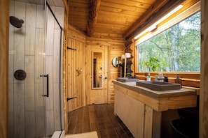 A wooden cabin bathroom with a shower, two sinks, a mirror, and a window with a view of trees.