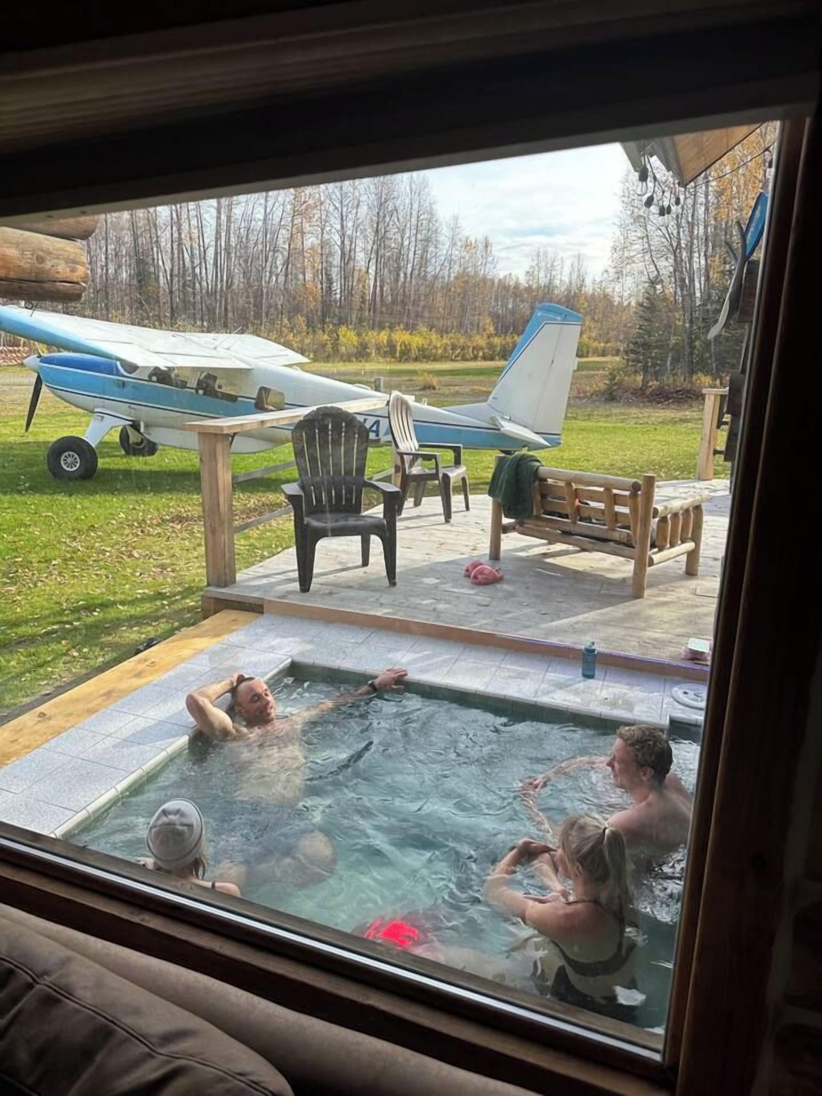 Un jacuzzi où se trouvent trois personnes, une terrasse en bois, une chaise et, au loin, un petit avion.