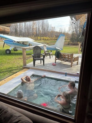 A hot tub with three people, a wooden deck, a chair, and a small airplane in the background.