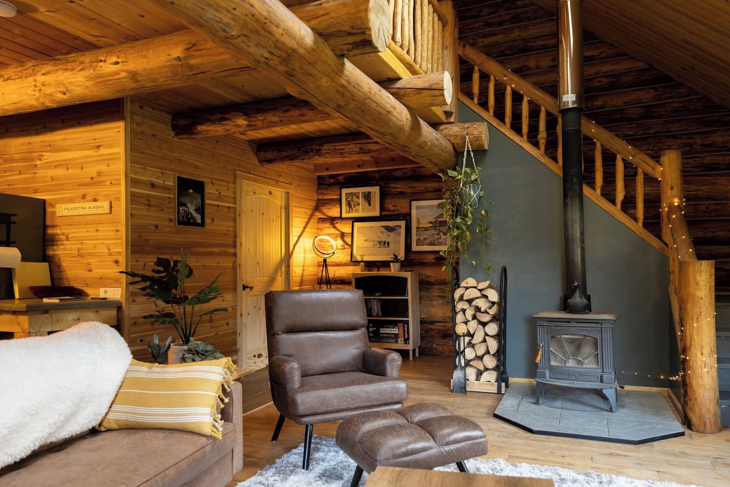 A cozy cabin interior with a wooden staircase, a leather armchair, a fireplace, and a sign reading 'Talkeetna Alaska'.