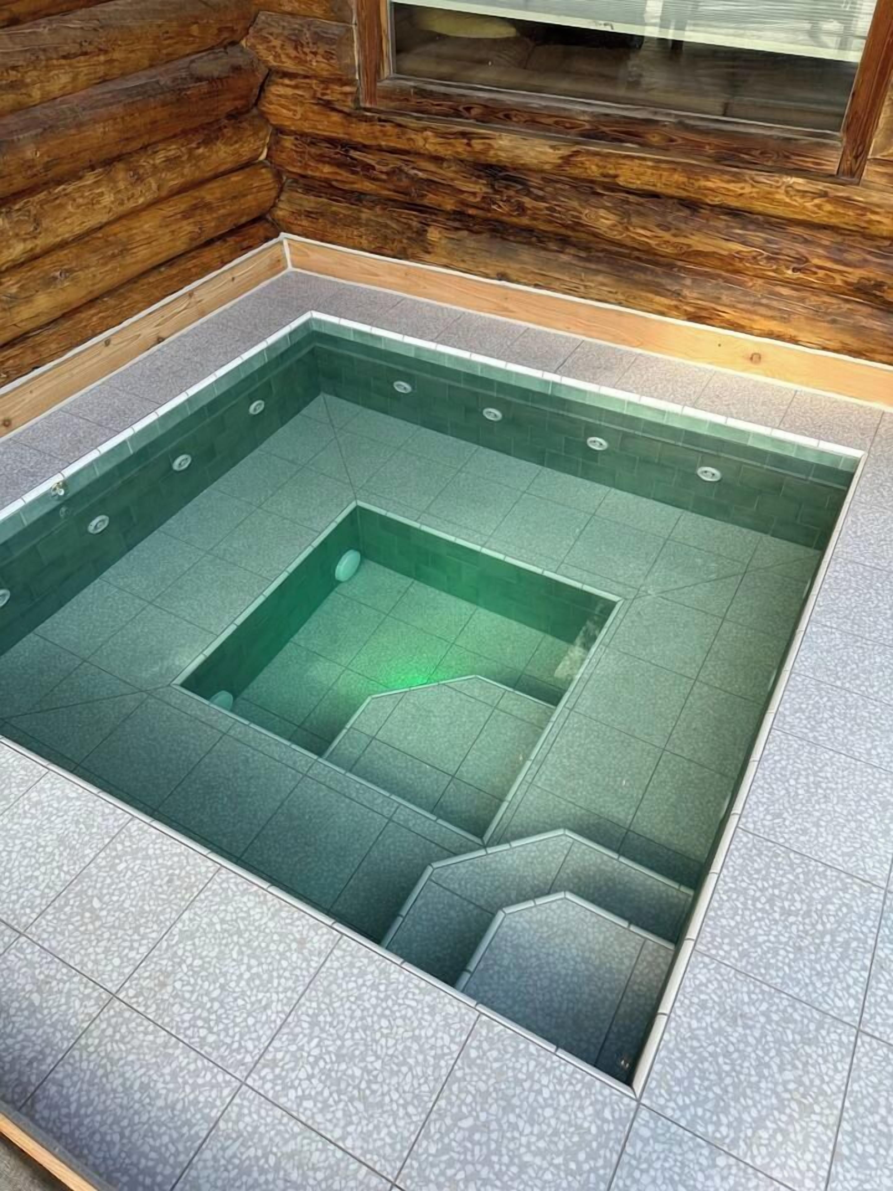 A square-shaped hot tub with a tiled floor and a wooden cabin wall in the background.