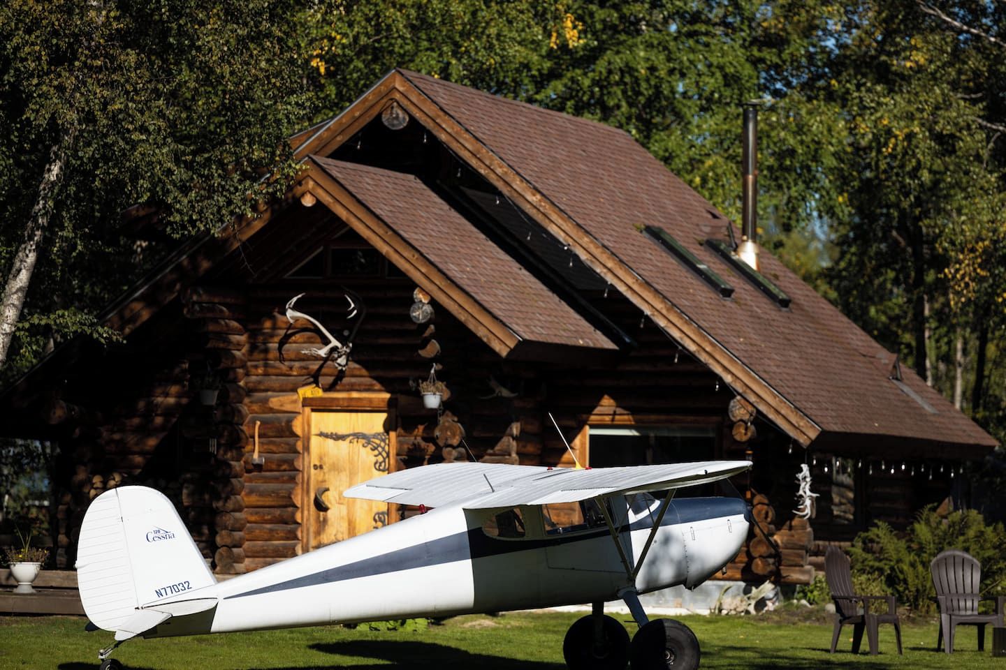 A small white airplane parked in front of a rustic log cabin with a sloped roof.