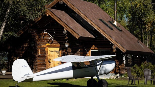 A small white airplane parked in front of a rustic log cabin with a sloped roof.