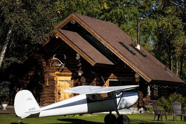 A small white airplane parked in front of a rustic log cabin with a sloped roof.