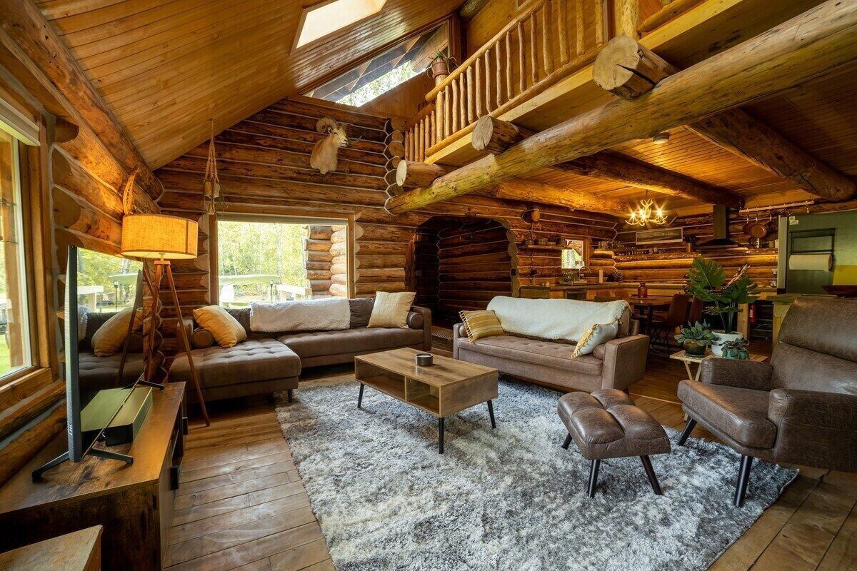 A log cabin interior with a wooden ceiling, a staircase, a sofa set, a coffee table, and a fireplace.