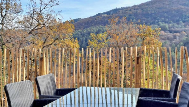 Una mesa y sillas frente a una valla de madera, con vista a árboles otoñales y colinas al fondo.