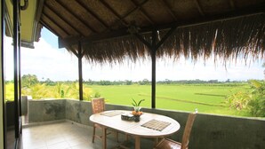 A covered outdoor area with a table and chairs, a thatched roof, and a view of a green field. - SUARA SIDHI Villa Ubud Bali (Ubud)