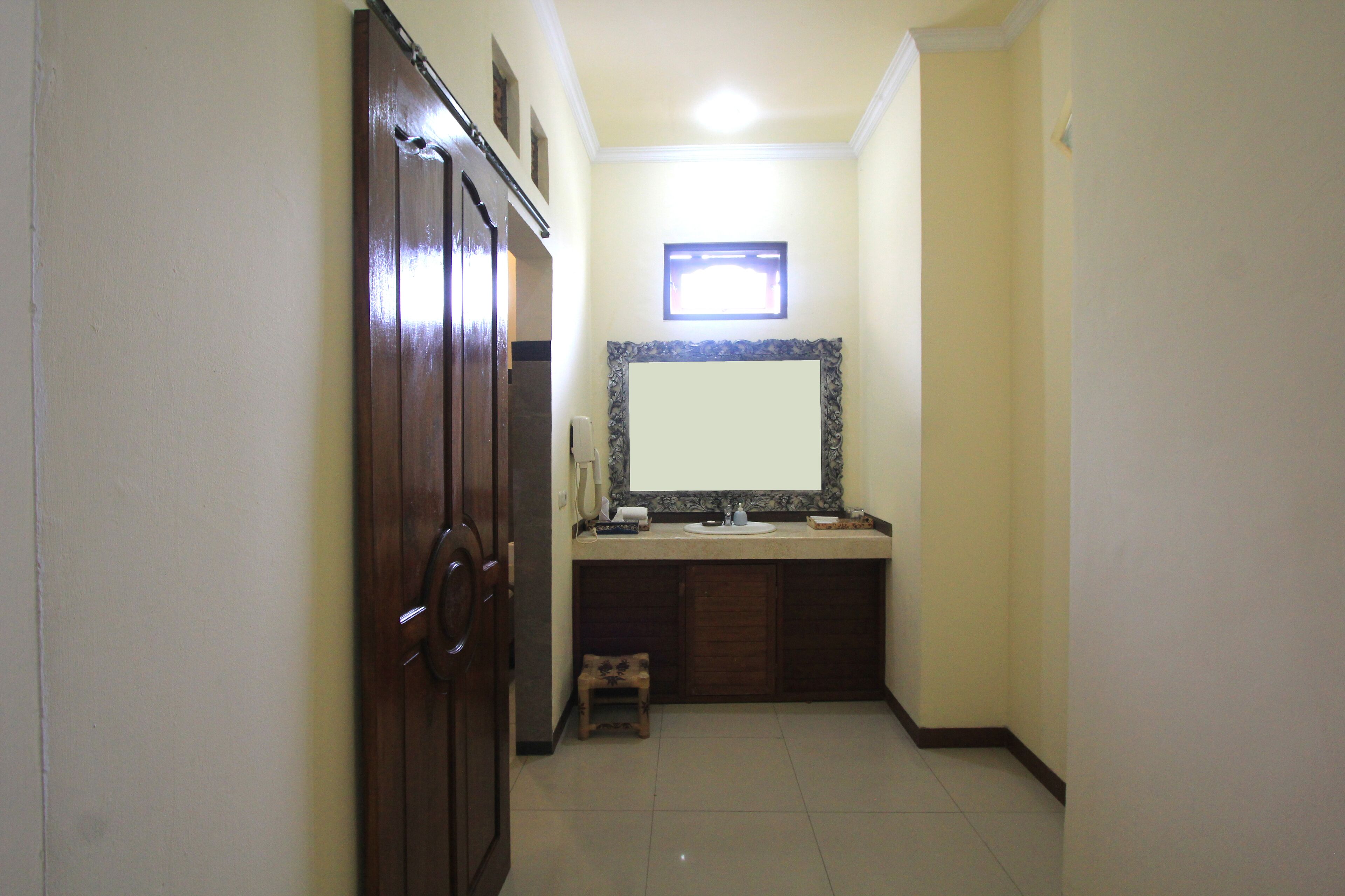 A narrow hallway leading to a bathroom with a wooden door, a small stool, a sink, and a mirror with a decorative frame.