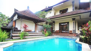 A swimming pool with a tiled deck, surrounded by plants and a building with a tiled roof and columns. - SUARA SIDHI Villa Ubud Bali (Ubud)