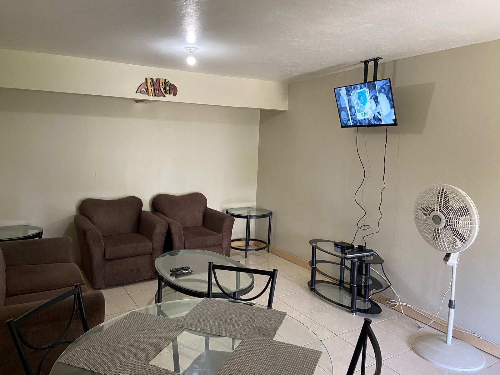 A living room with two brown sofas, a glass coffee table, a TV mounted on the wall, and a standing fan.