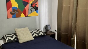 Un dormitorio con cama, ventilador de techo, un cuadro, mesita de noche y cortinas.