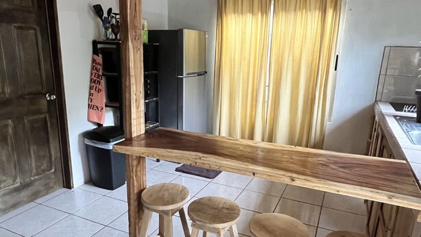 Una cocina con un taburete de madera bajo una viga de madera, suelo de baldosas y una ventana con cortinas.
