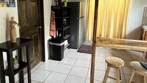Una habitación amplia con suelo de baldosas, techo de madera, una puerta, un estante negro con varios objetos, un taburete pequeño y un banco.