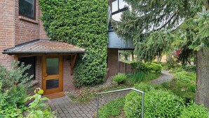 A brick building with a wooden door, a brick pathway, and a garden with various plants and trees. - Haus Ersfeld – Garden Apartment with Private Terrace, Shared Garden, and Wi-Fi (Bad Teinach-Zavelstein)