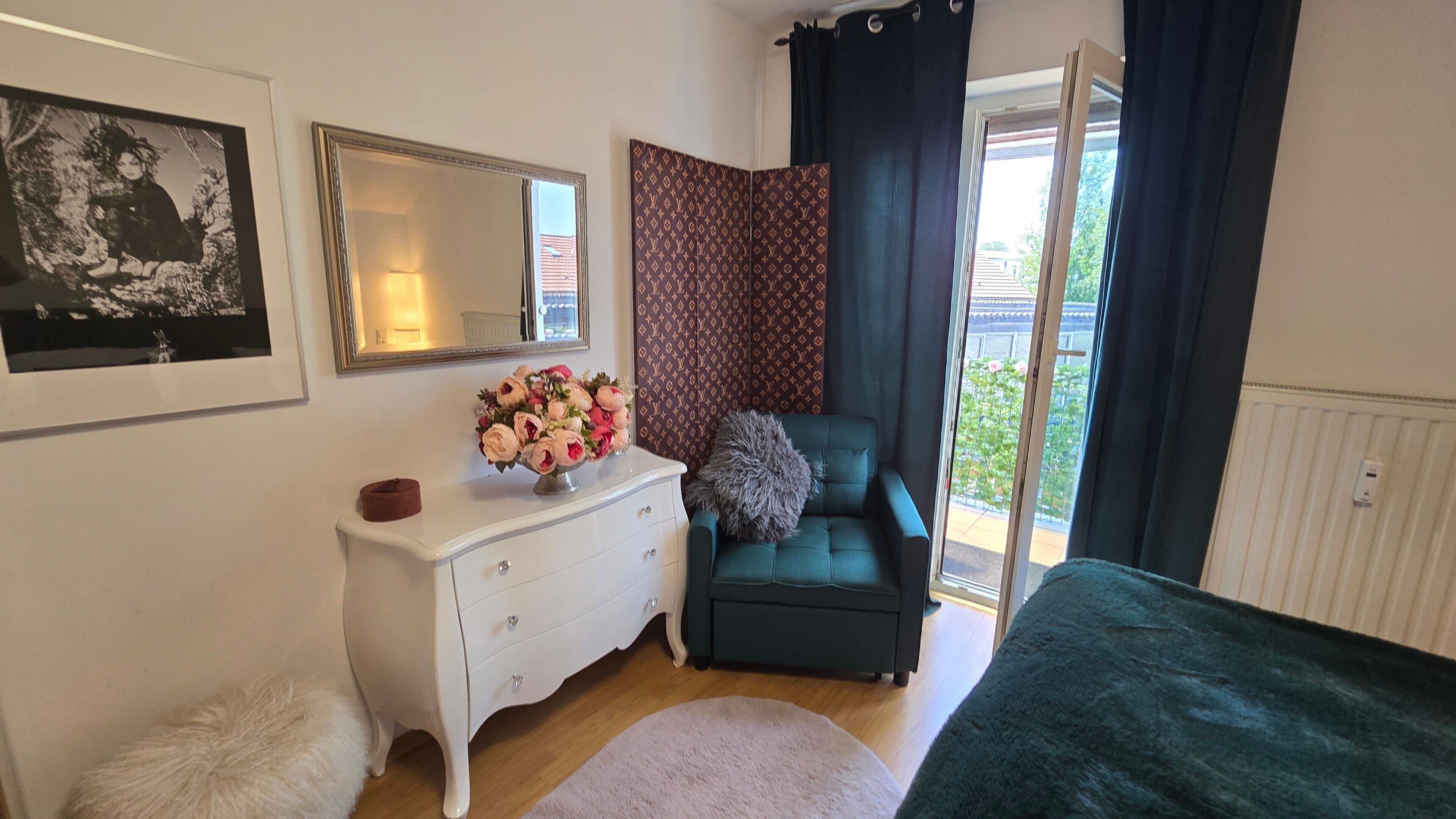 Une chambre à coucher avec une commode, un canapé vert, un lit, un miroir et une fenêtre avec des rideaux.