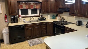 Une cuisine avec des meubles de couleur bois foncé, un plan de travail blanc, une cuisinière, un évier et une fenêtre agrémentée d’une lambrequin décorative.